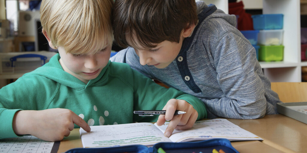 Zwei Kinder beugen sich gemeinsam über ein Arbeitsheft und lesen.