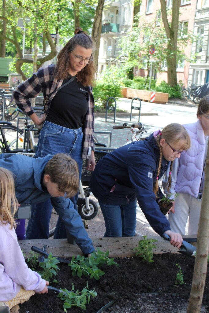 Eine Erwachsene und mehrere Kinder pflanzen Setzlinge in ein Hochbeet.