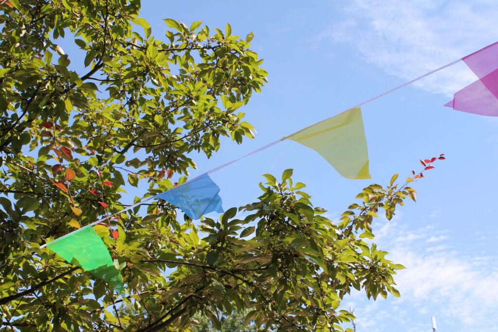 Bunte Wimpelkette zwischen grünen Zweigen vor blauem Himmel.