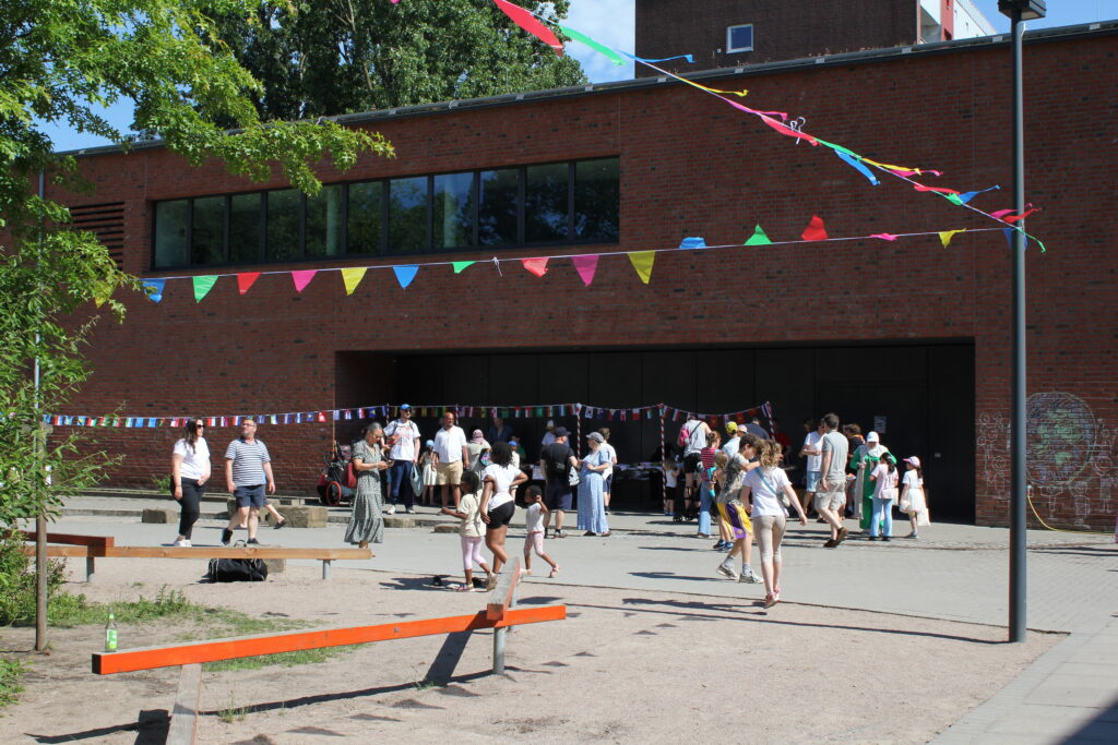 Sommerfest auf dem Schulhof: viele Menschen unter bunten Wimpelketten vor der Sporthalle.