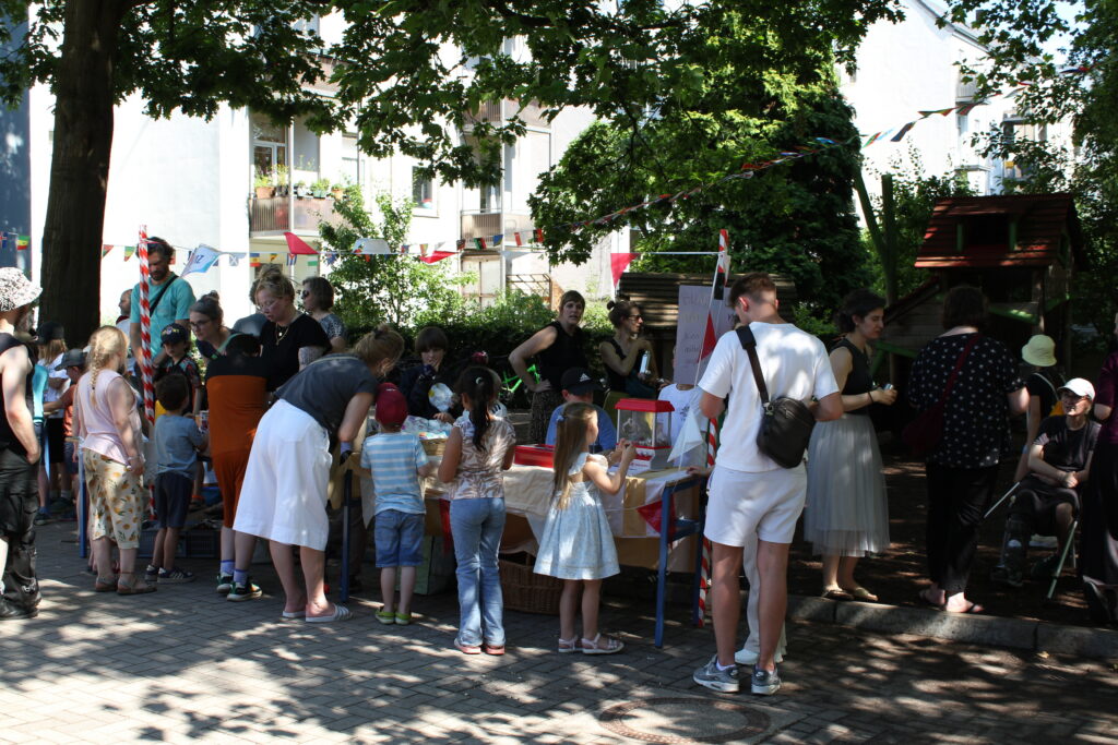 Besucherinnen und Besucher drängen sich beim Sommerfest um einen Stand im Schatten der Bäume.