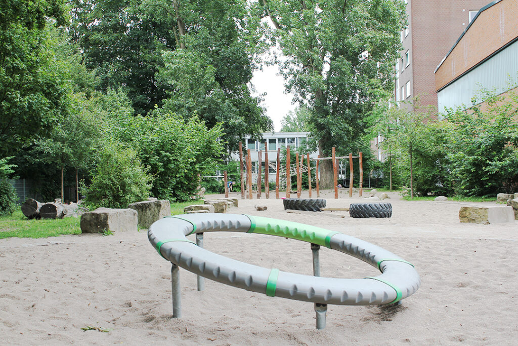 Schulhof mit einem runden Klettergerät im Sand, dahinter Bäume und ein Holzklettergerüst.