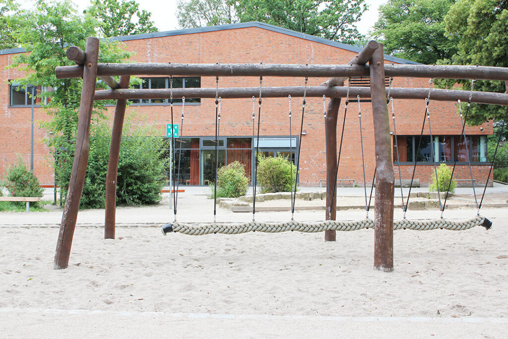 Holzschaukelgerüst mit Netzschaukel auf dem sandigen Schulhof, dahinter die Sporthalle.