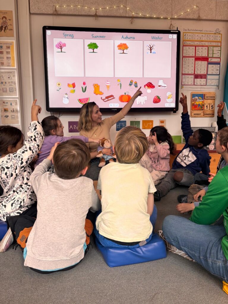 Kinder sitzen vor einer digitalen Tafel mit Bildern zu den Jahreszeiten.