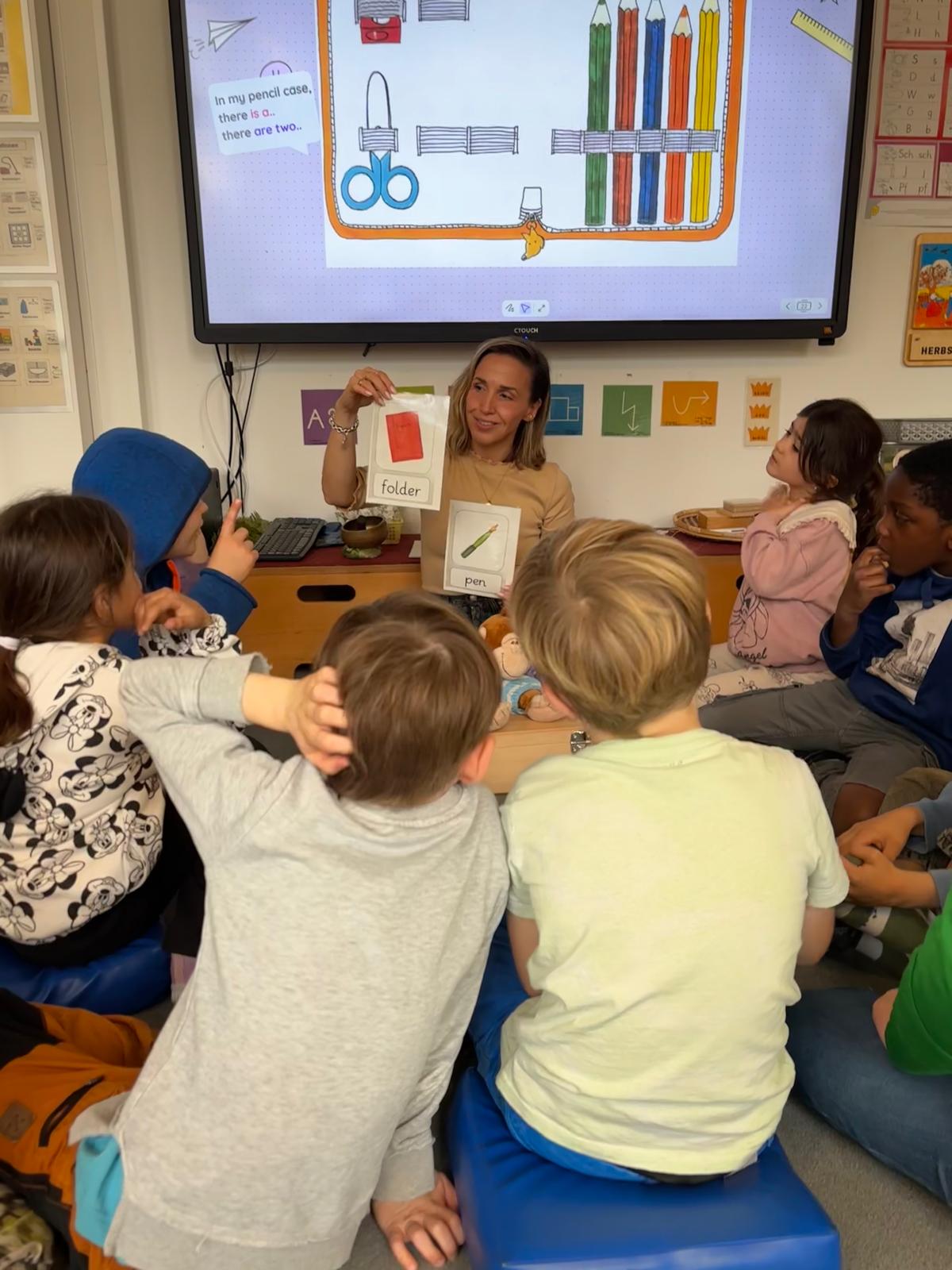 Kinder sitzen im Kreis vor einer digitalen Tafel, eine Lehrerin erklärt.