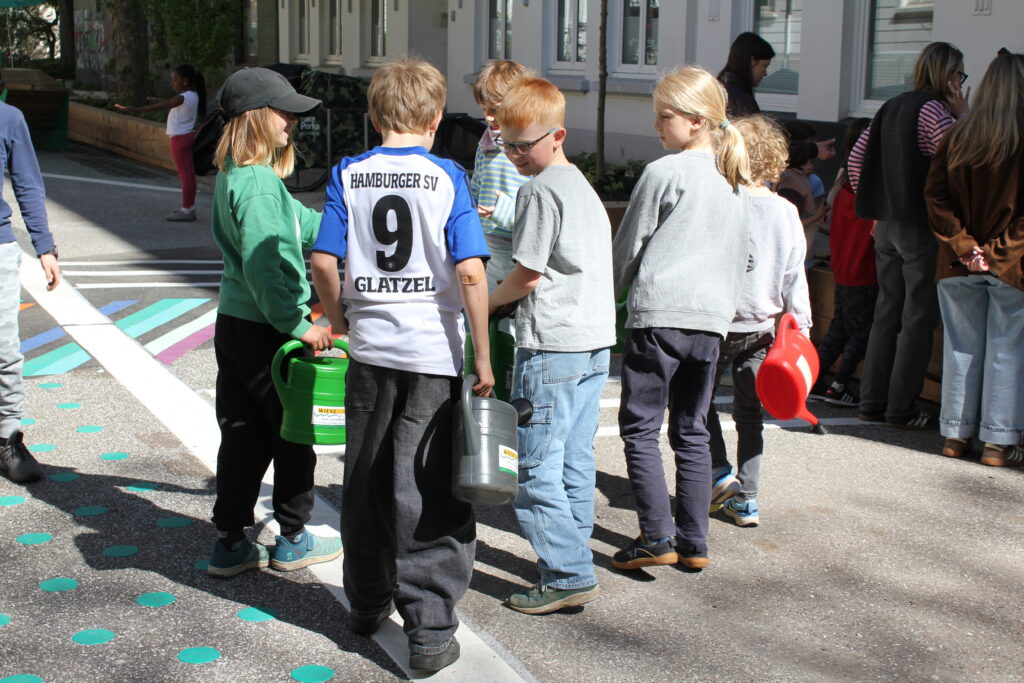 Kinder mit Gießkannen stehen in einer Reihe auf der Spielstraße.