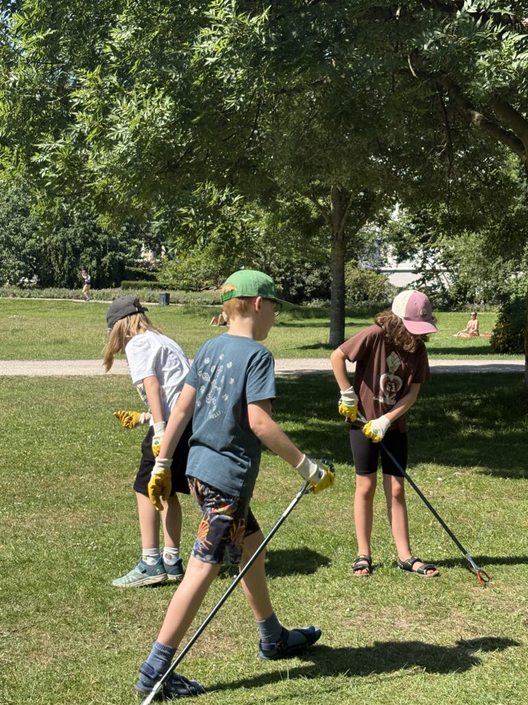 Drei Kinder mit Handschuhen und Greifzangen sammeln Müll auf der Wiese