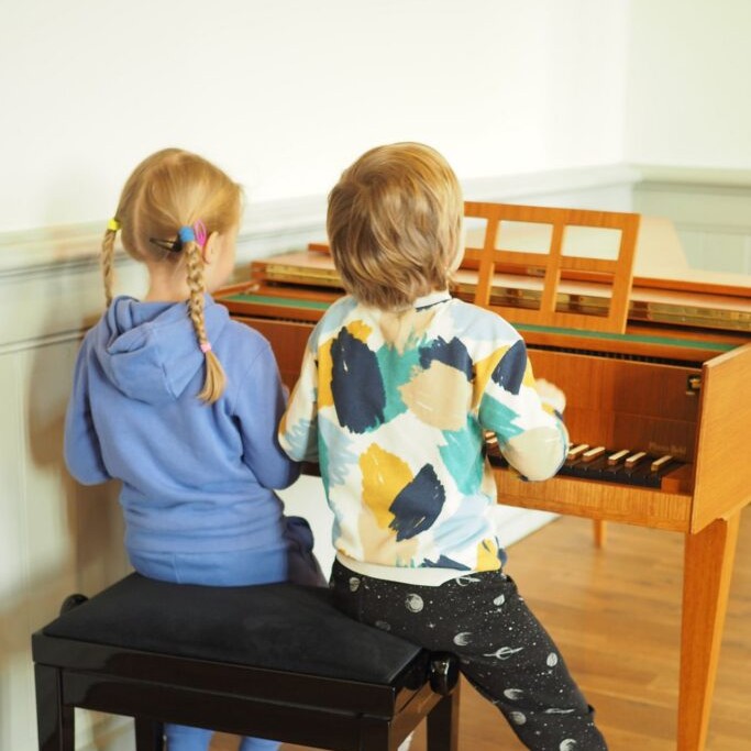 Zwei Kinder sitzen nebeneinander am Klavier, von hinten fotografiert.