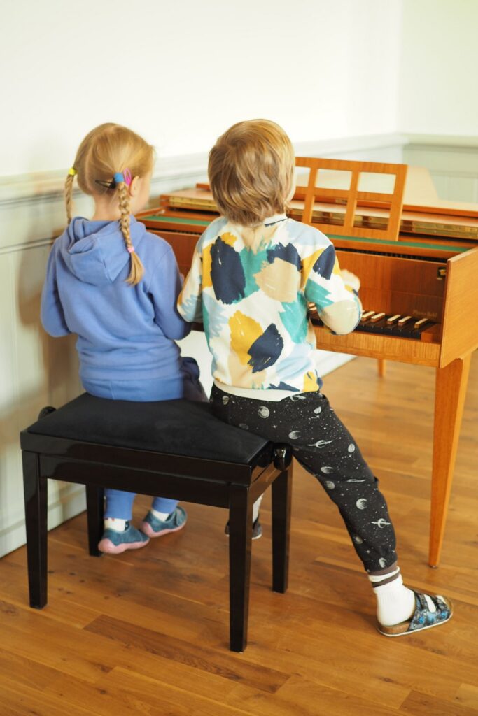 Zwei Kinder sitzen nebeneinander am Klavier, von hinten fotografiert.