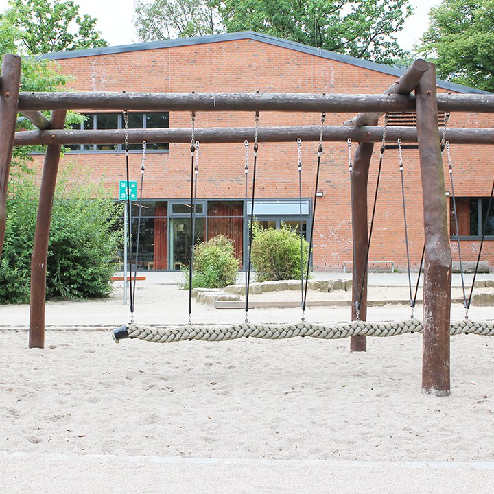 Holzschaukelgerüst mit Netzschaukel auf dem sandigen Schulhof, dahinter die Sporthalle.
