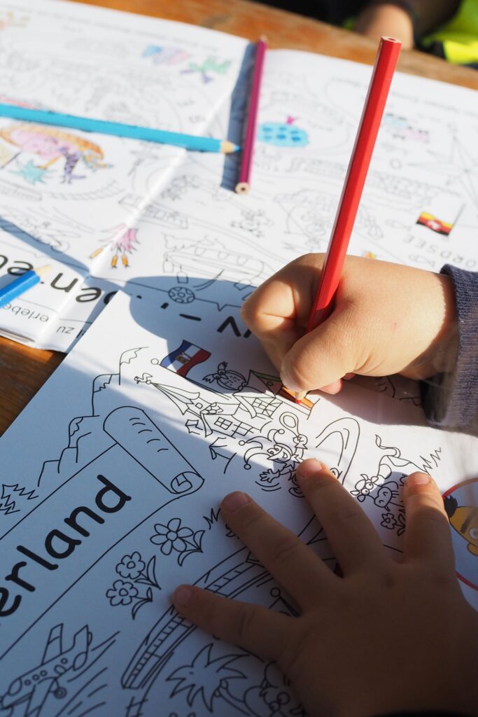Eine Kinderhand malt mit Buntstift ein Bild in einem Heft aus.