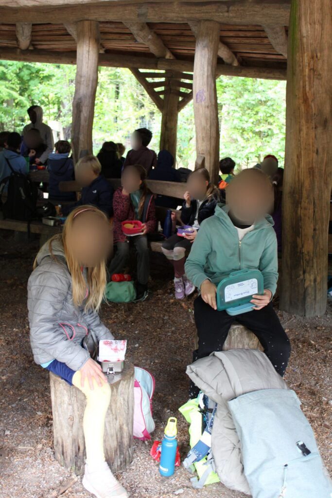 Kinder sitzen unter einem Holzunterstand im Wald und frühstücken.
