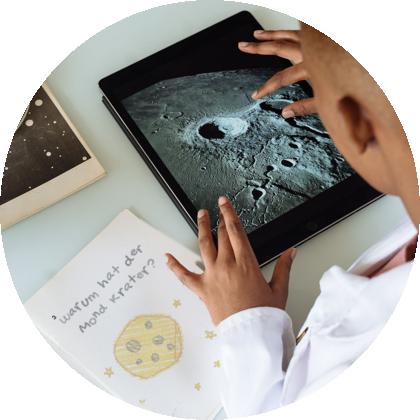 Kinderhände auf einem Tablet mit einem Foto der Mondoberfläche, daneben ein Blatt mit der Frage „Warum hat der Mond Krater?“.